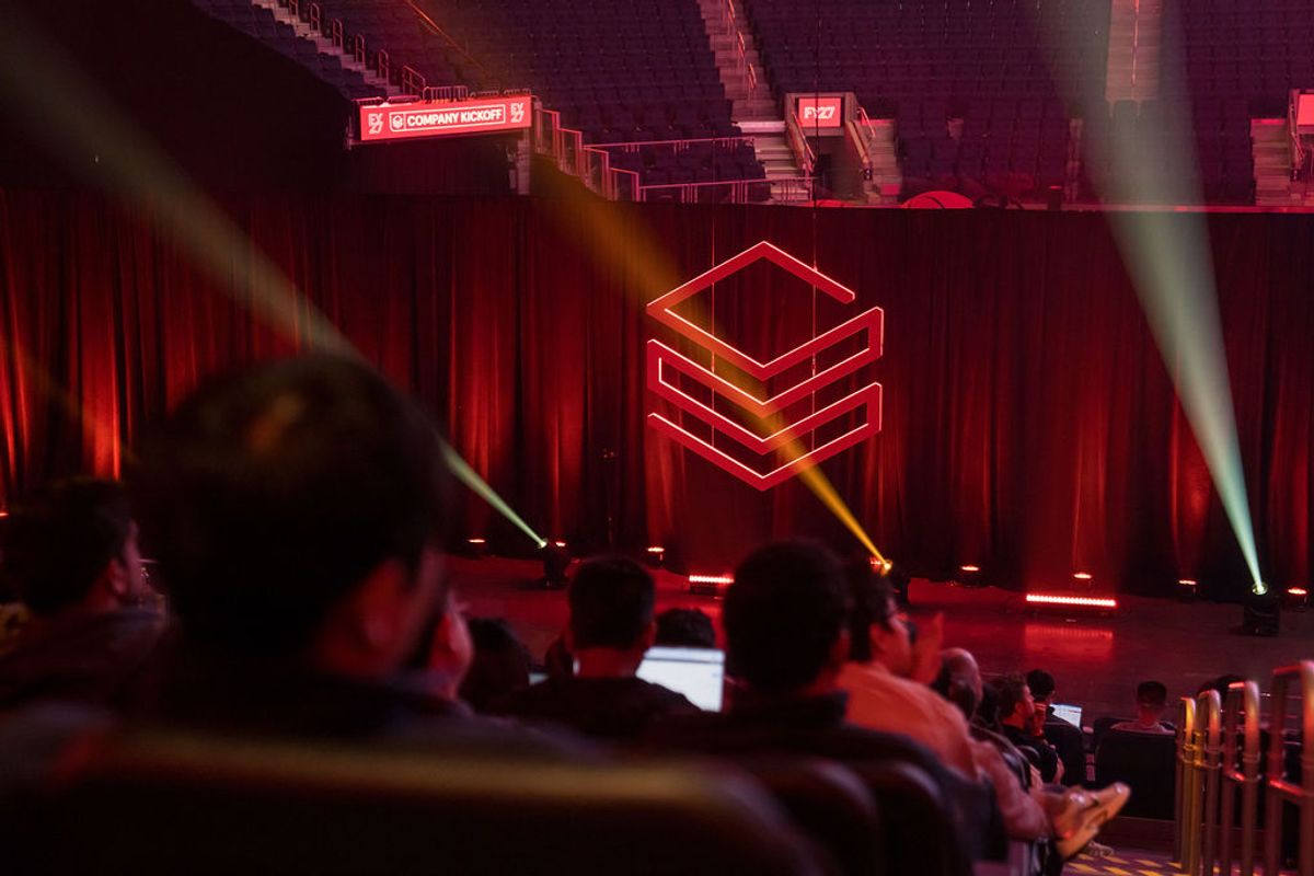 Conference attendees watch a presentation with red geometric logo and dramatic lighting at an arena venue