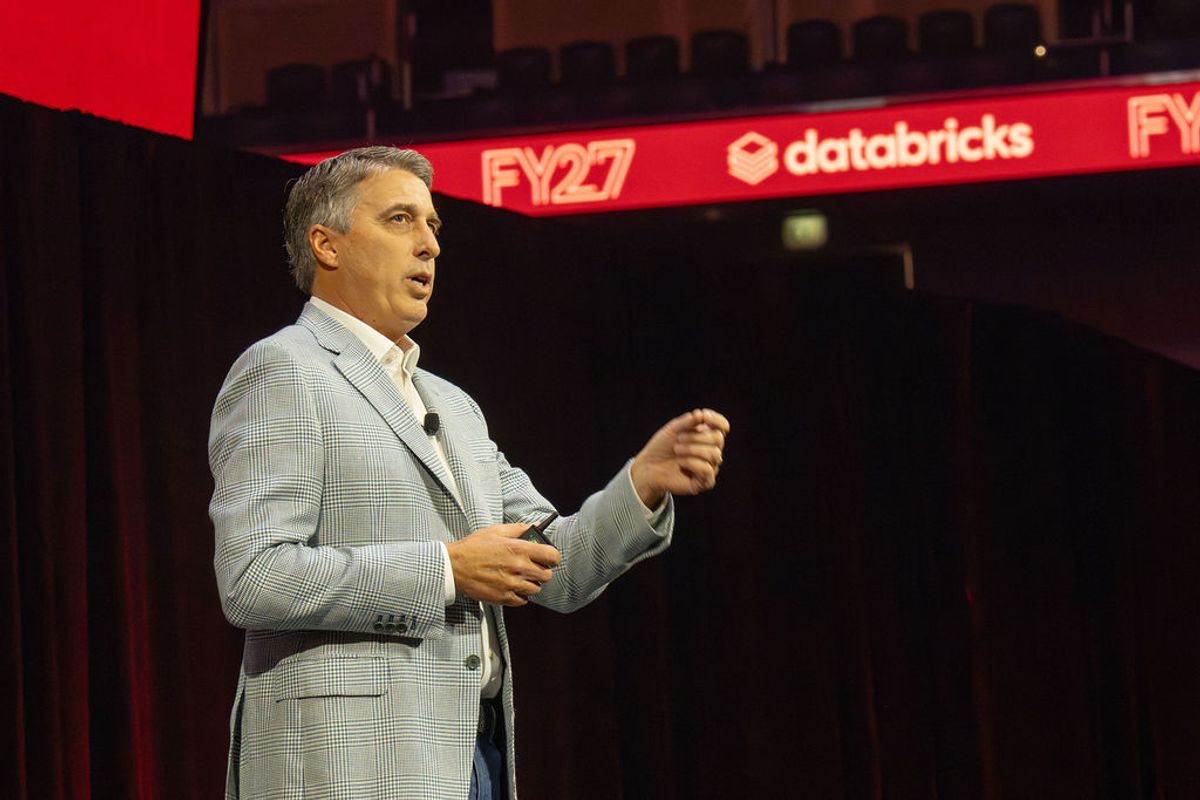 Executive in gray suit presenting on stage at conference with red Databricks and FY27 signage visible