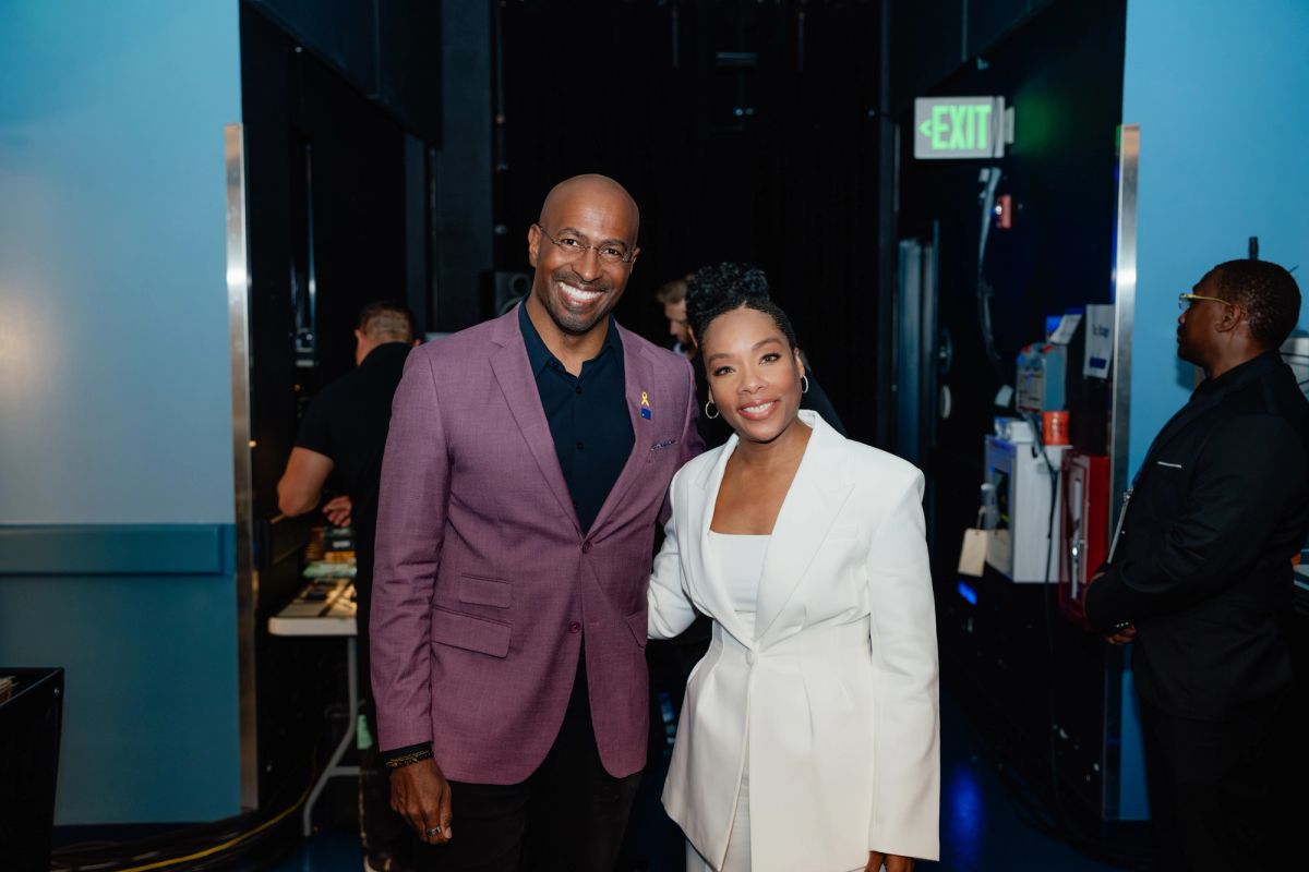 Two people pose together backstage at an event, one wearing a purple blazer and one in a white outfit.