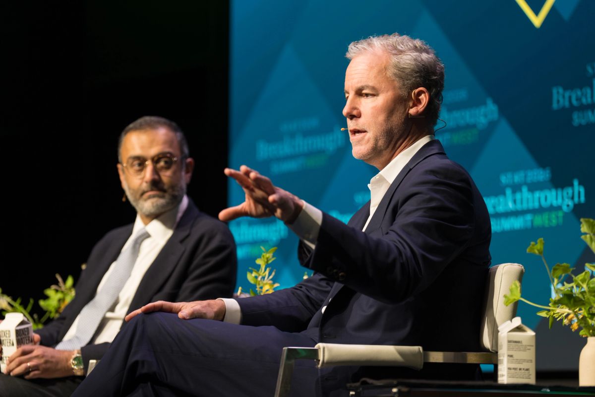 Two men in business suits sitting on stage during a panel discussion, one gesturing while speaking