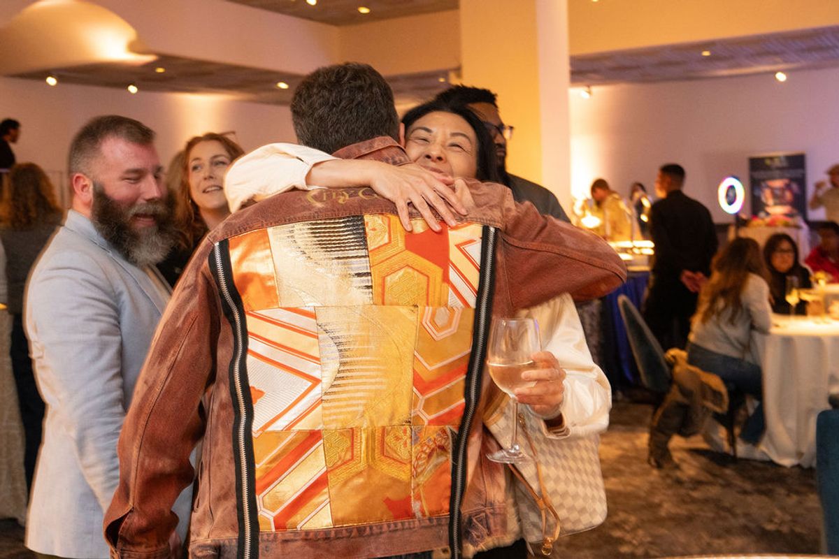 Two people embrace warmly at a networking reception while other attendees socialize in the background.