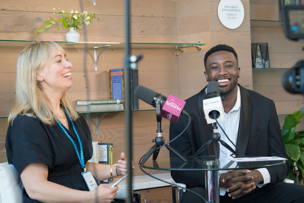 Two speakers laugh during a podcast recording, sitting at microphones with shelving and plants in background.