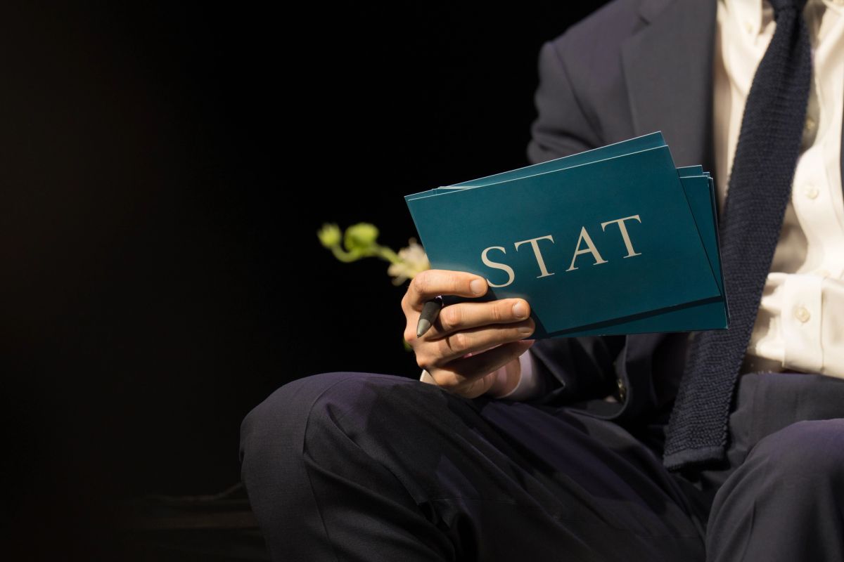 Speaker in suit holding teal folders with "STAT" text during conference presentation