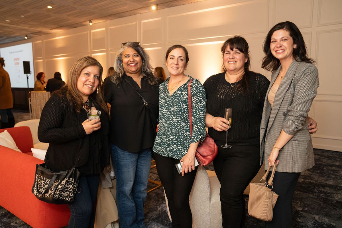 Five women networking and posing together with drinks at a professional reception event indoors.