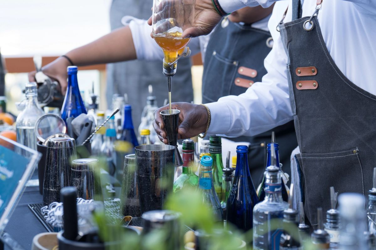 Bartender in apron pours golden liquid into cocktail shaker at outdoor bar setup with bottles and equipment