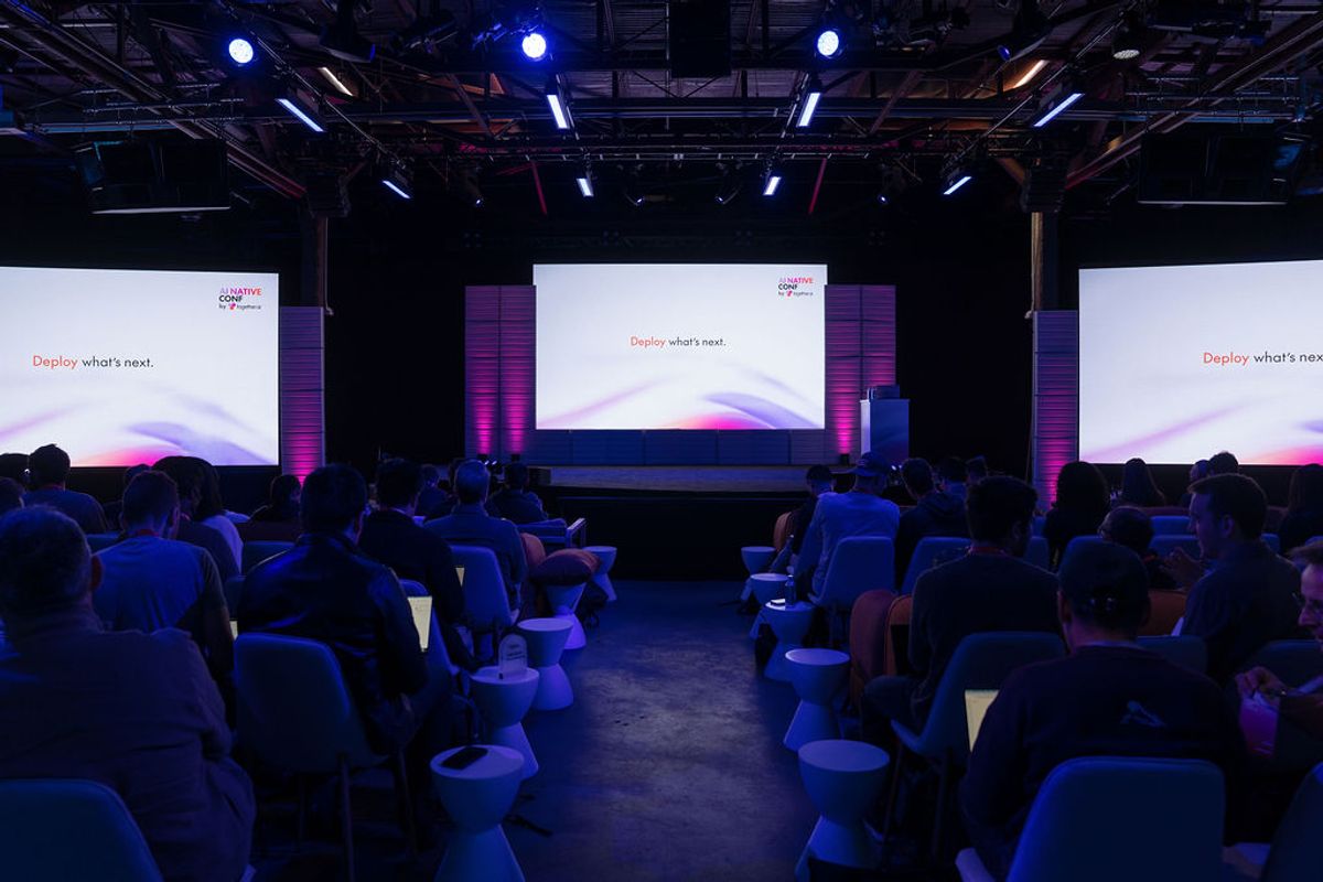 Conference attendees seated in chairs facing a large screen displaying "Deploy what's next" with purple.