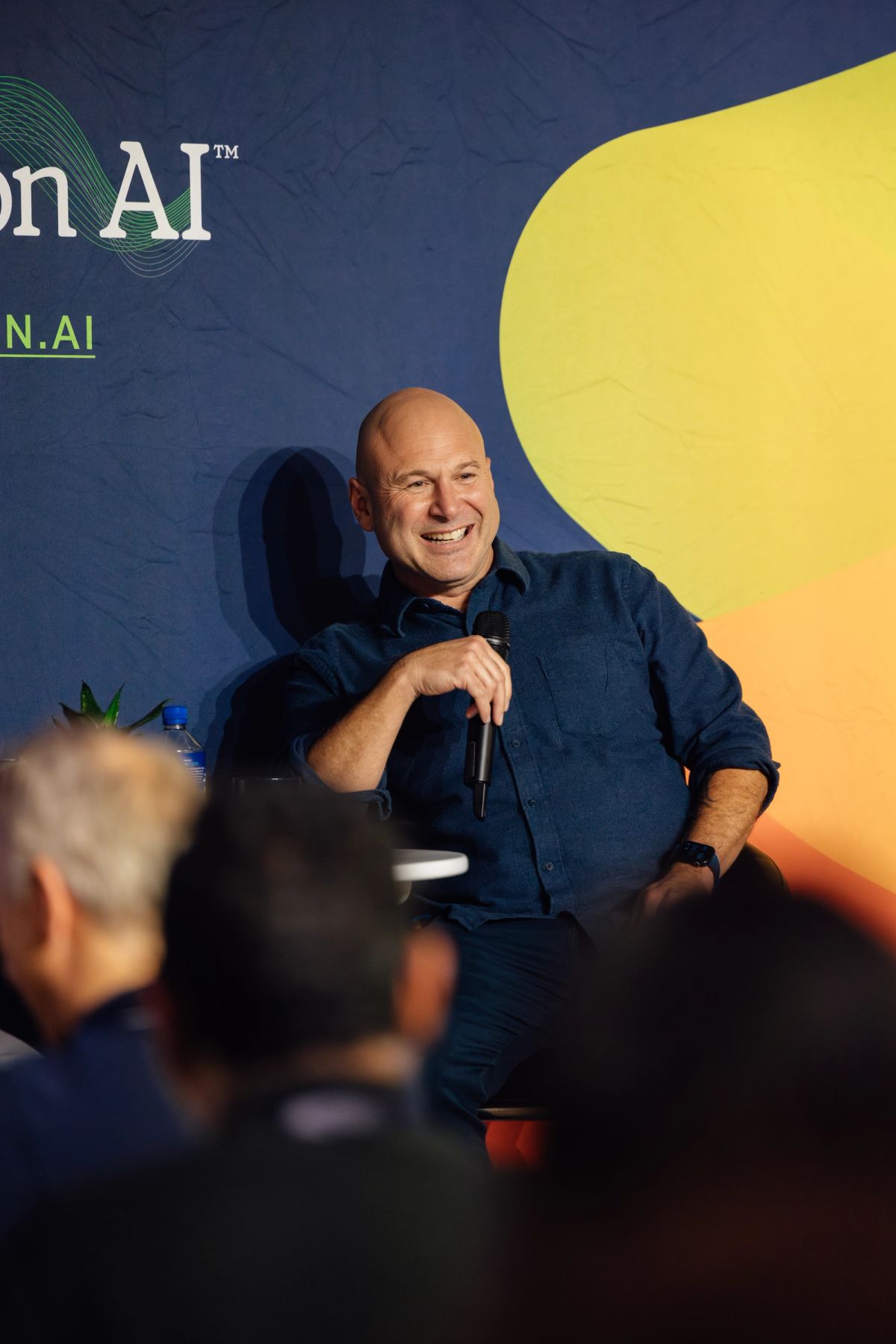 Smiling bald speaker in blue shirt holds microphone at conference, with AI-themed backdrop and audience.