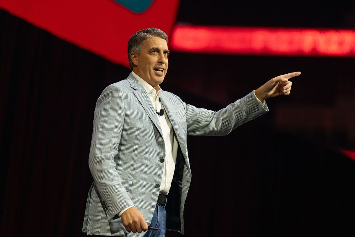 Speaker in gray suit gesturing while presenting on stage with red lighting in background
