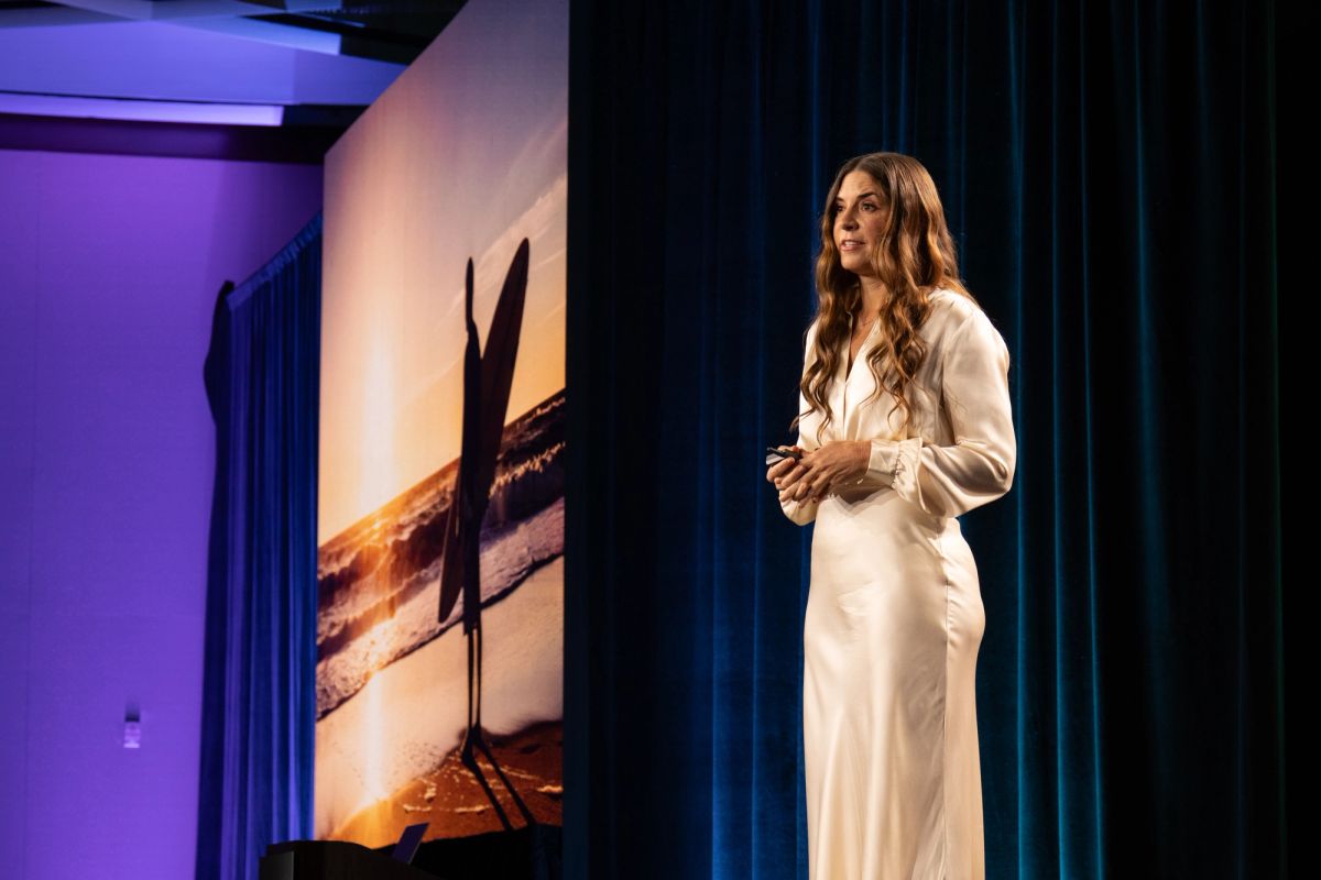 Woman in white dress presenting on stage beside a projected sunset beach photo.