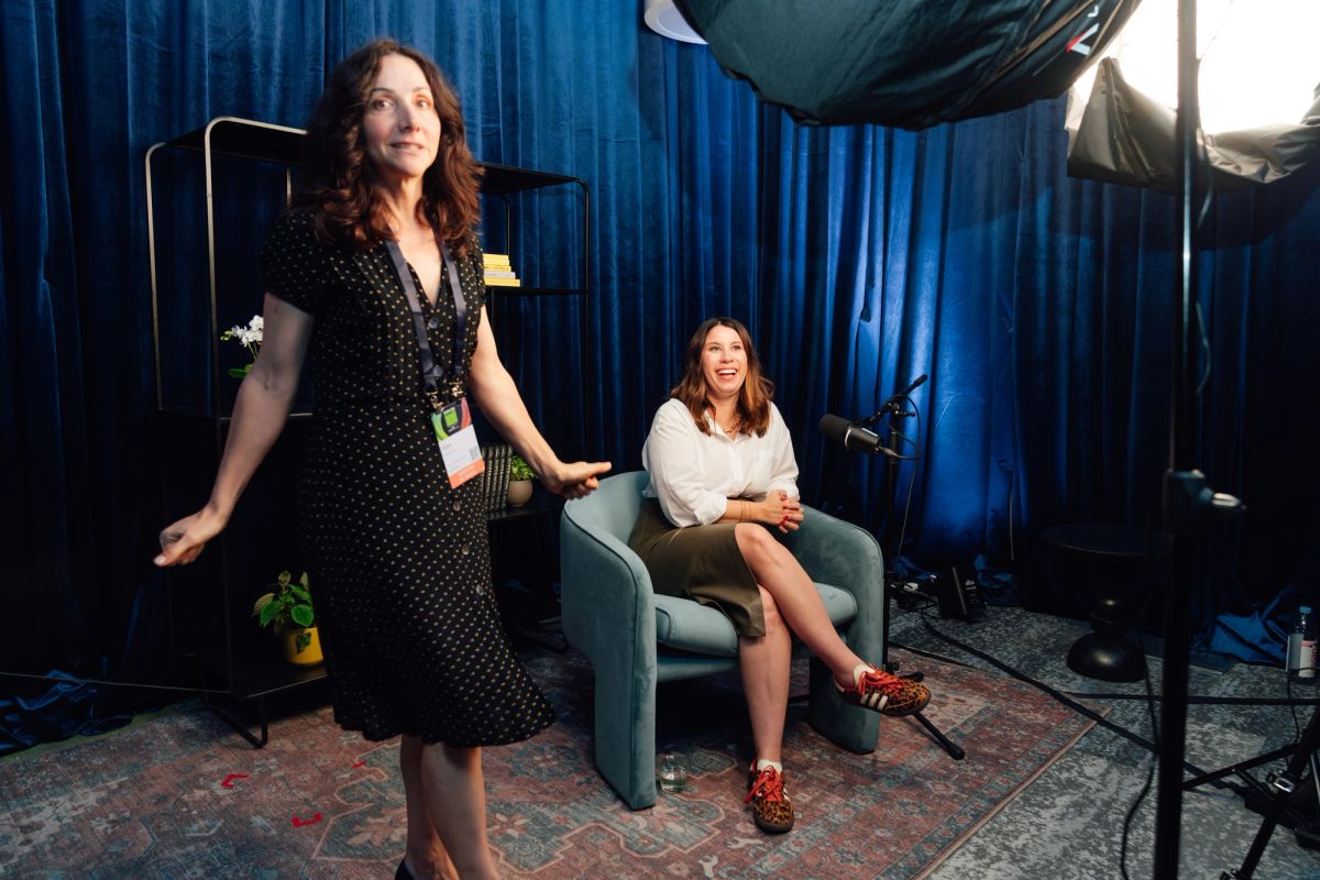 Two women in interview setup with blue backdrop, one standing in polka dot dress, one seated in chair
