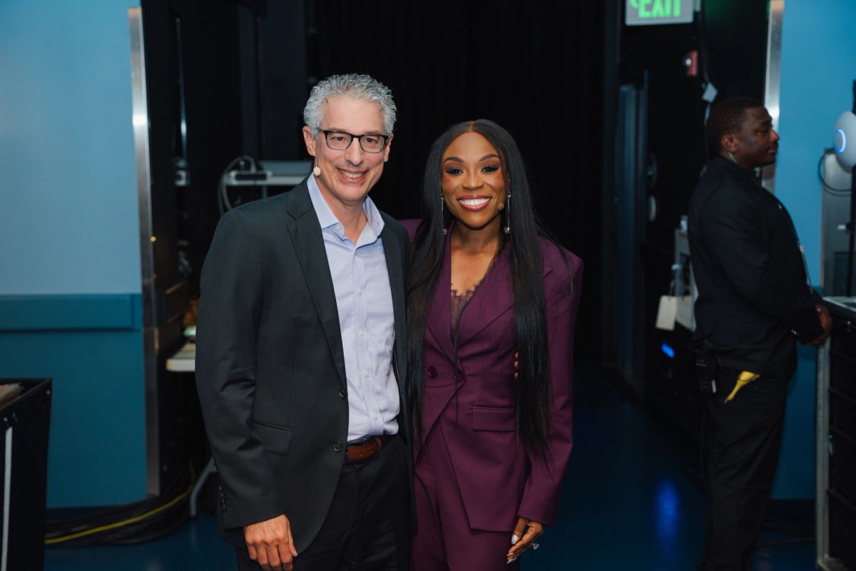 Two people posing together backstage at an event, one in a dark suit and one in a burgundy blazer.