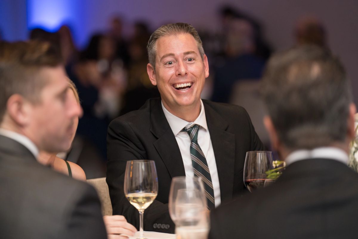 Man in dark suit laughing at dinner table with wine glasses, other attendees visible in background at formal.