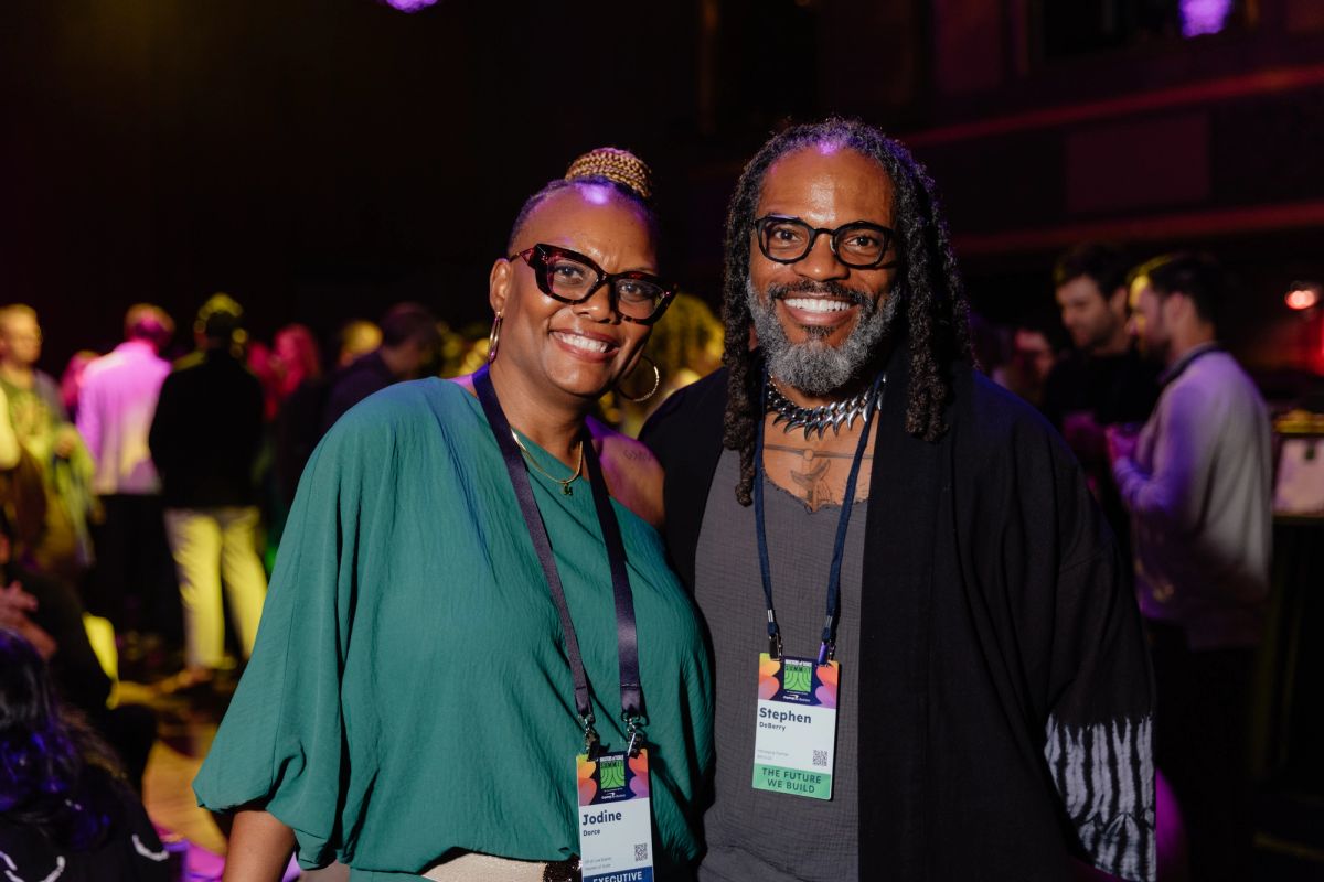 Two conference attendees with name badges smile together at an indoor networking event with purple lighting.