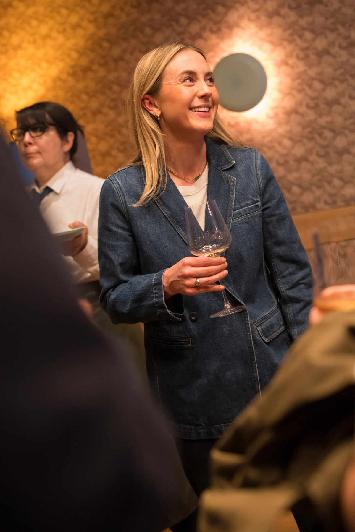 Woman in denim blazer holding wine glass smiles while looking up at panel discussion event with attendees in.