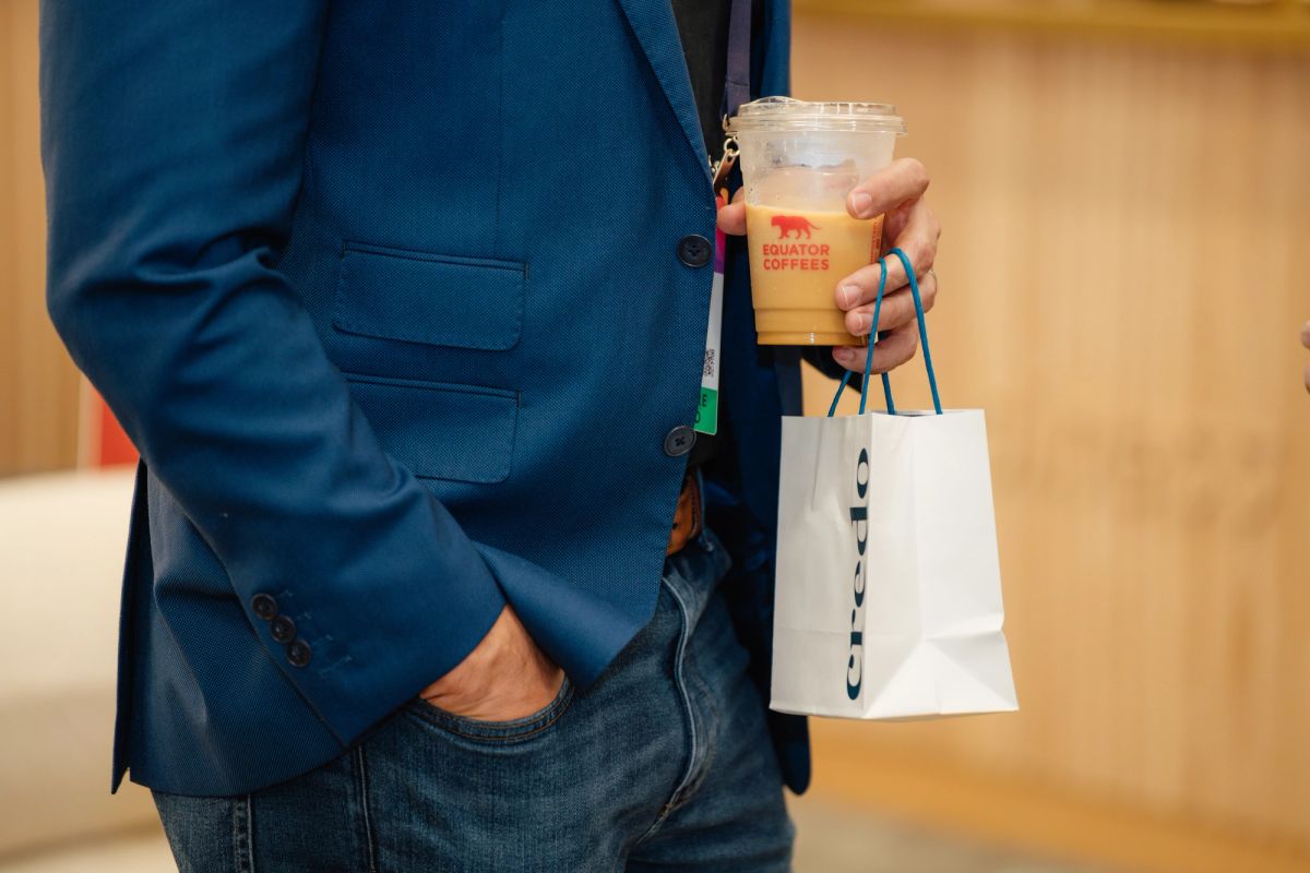 Person in blue blazer holding iced coffee and white shopping bag at indoor event