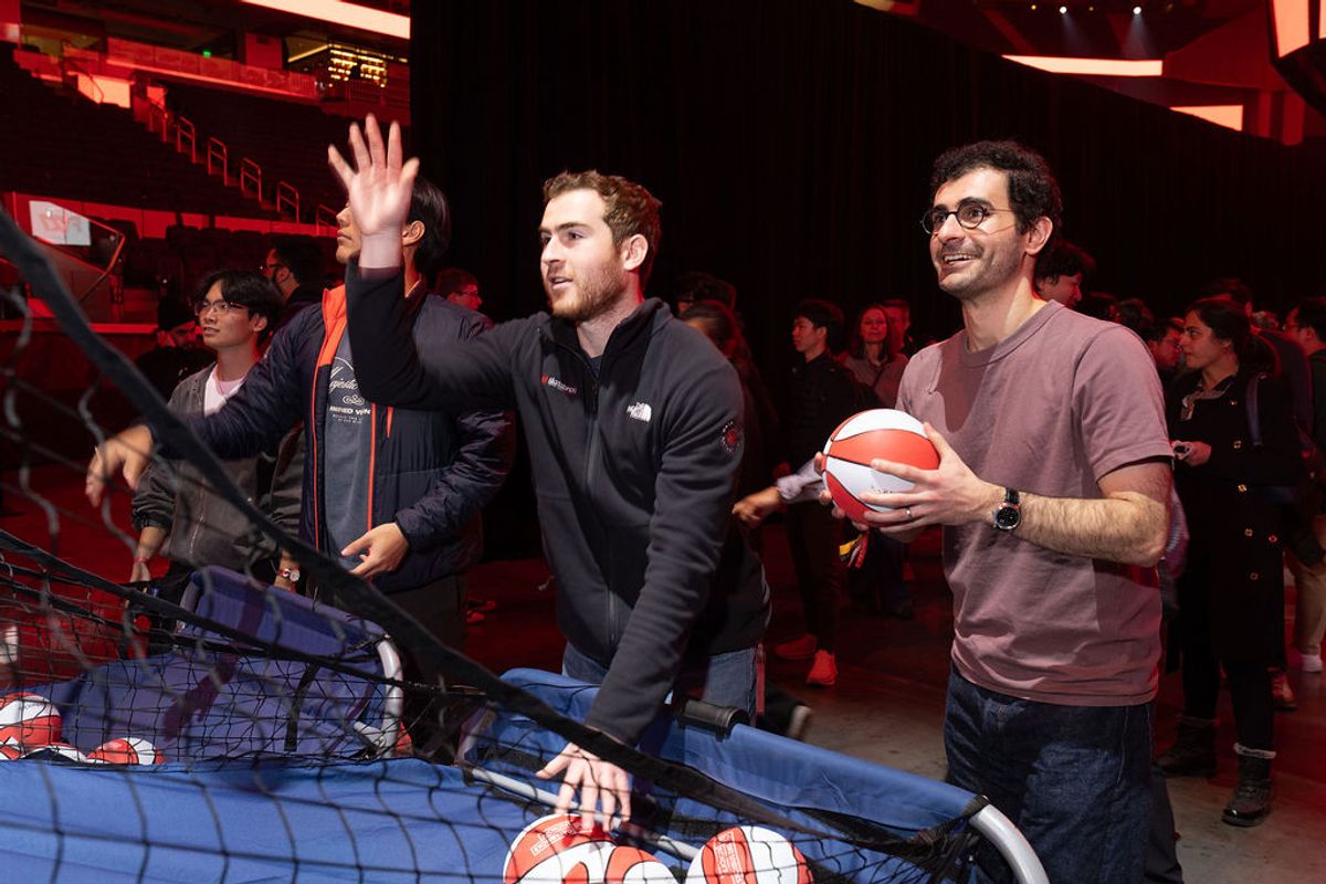 Two men at basketball shooting game, one waving and one holding basketball, with crowd in background