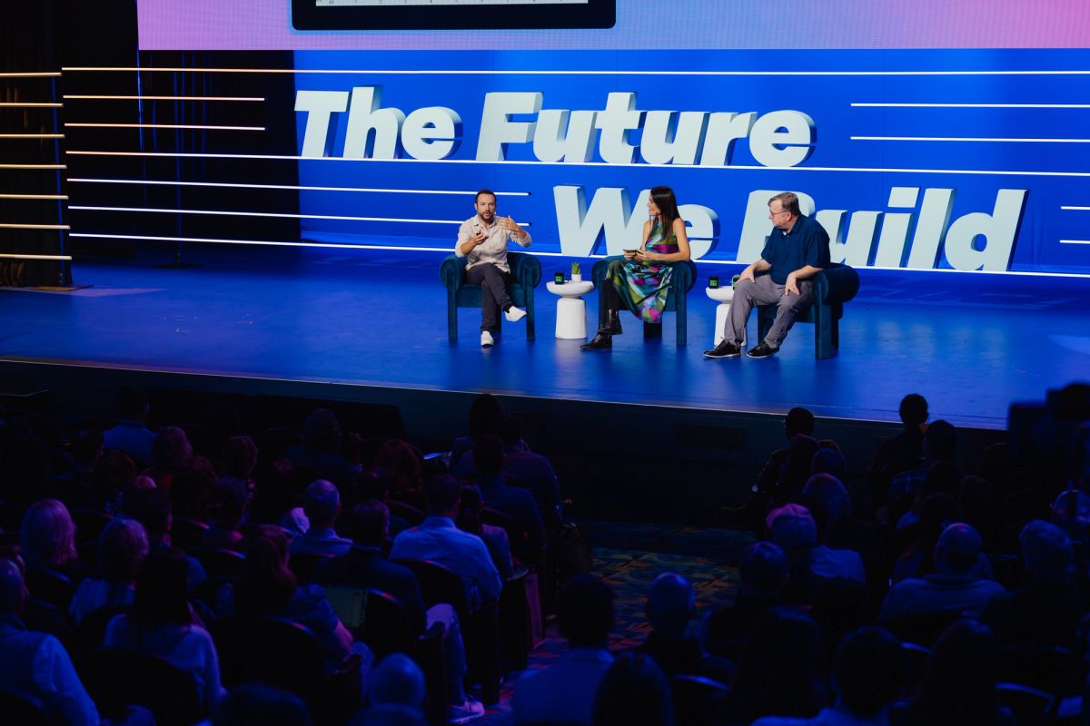 Three panelists sit in chairs on blue stage with "The Future We Build" backdrop before seated audience