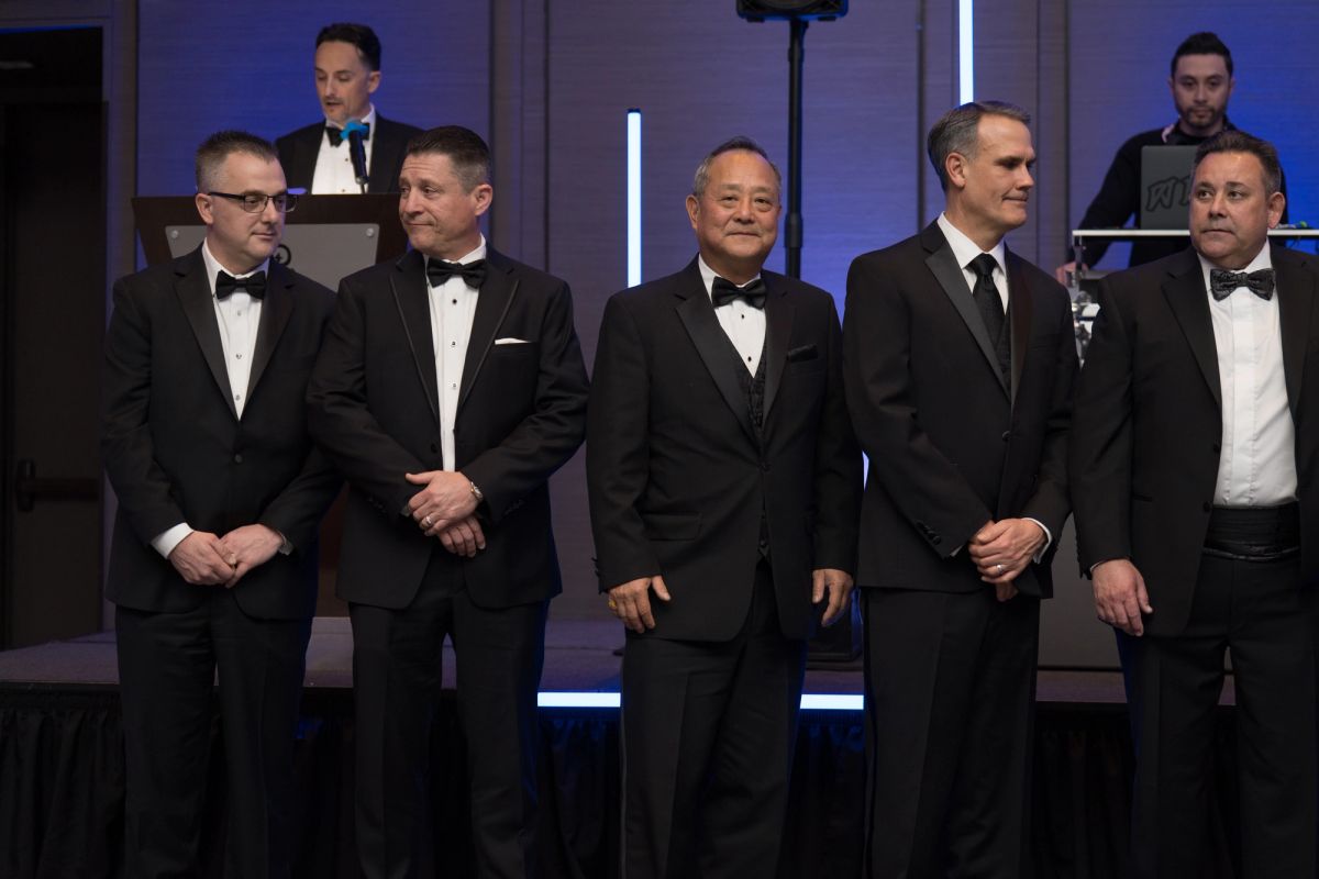 Five men in tuxedos standing together at formal event, with speaker at podium in background
