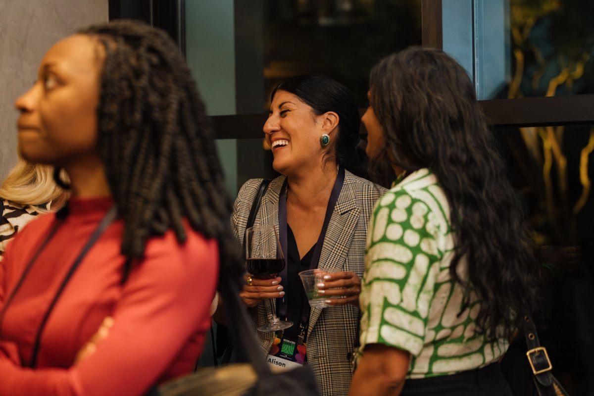 Three women networking at a conference, holding drinks and smiling while conversing in an indoor venue.