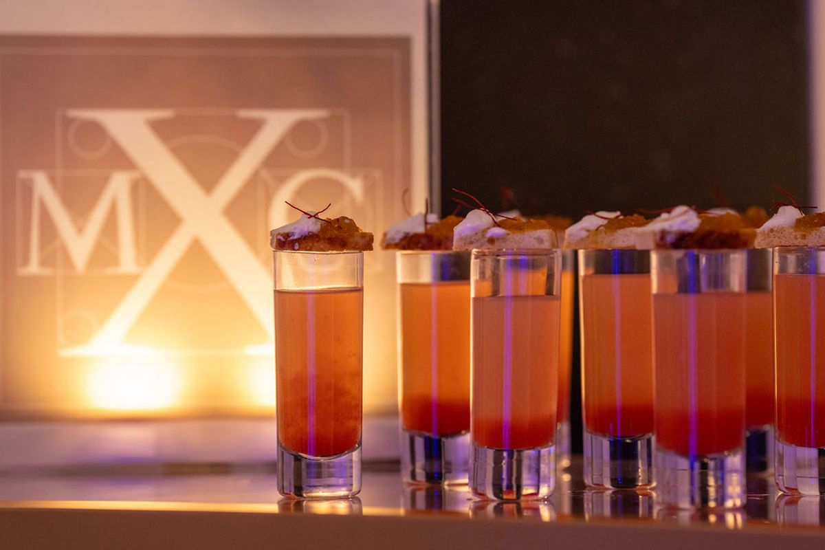 Row of orange cocktails with garnishes on a bar table with illuminated "MXC" signage in background