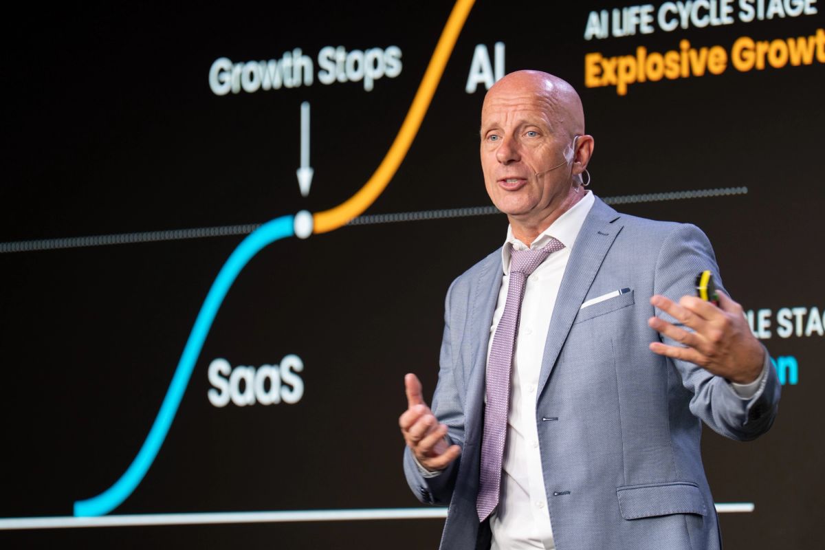 Business presenter in gray suit speaking at conference with AI life cycle growth chart displayed on screen.