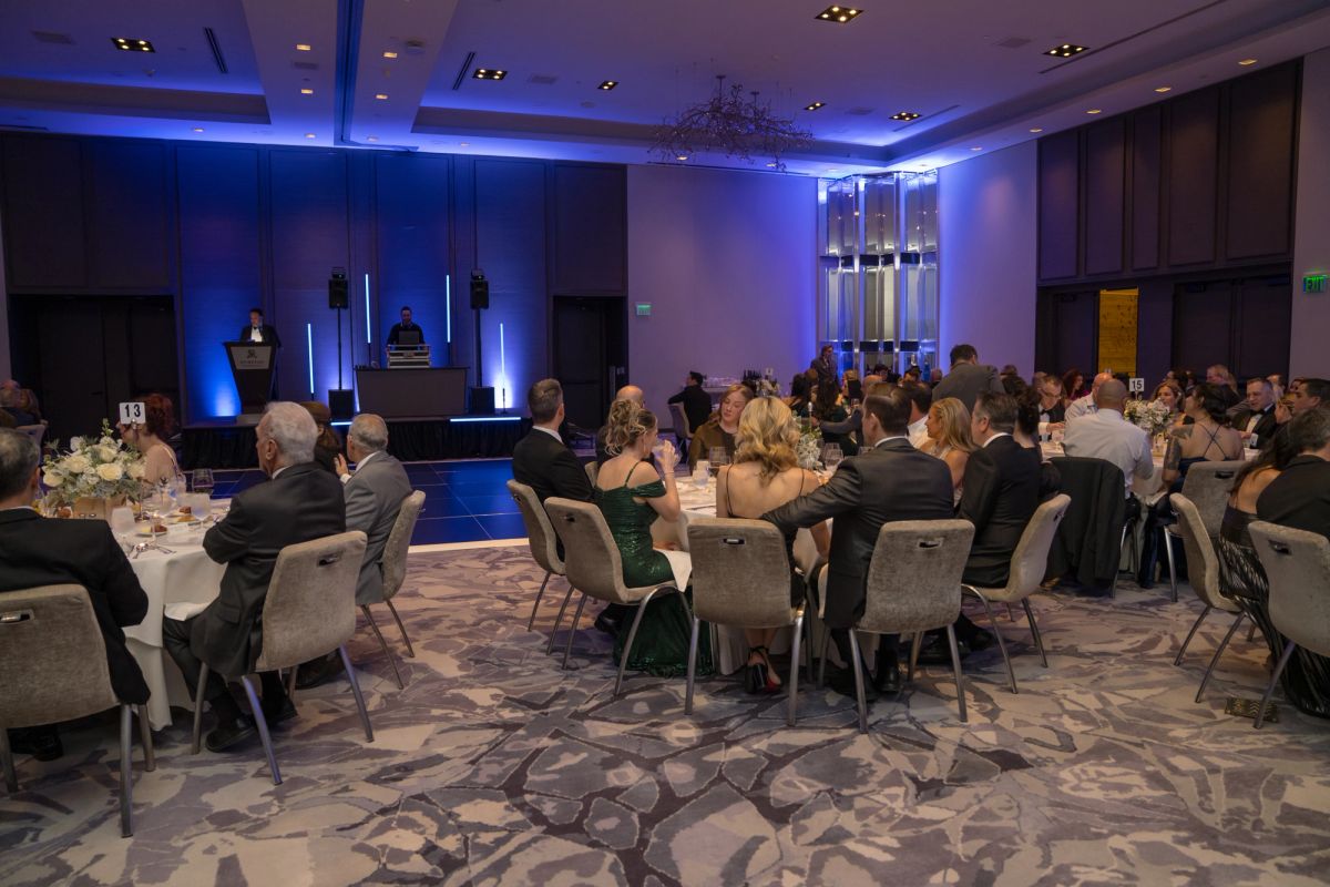 Formal gala dinner with attendees seated at round tables in an elegantly lit ballroom with stage backdrop.