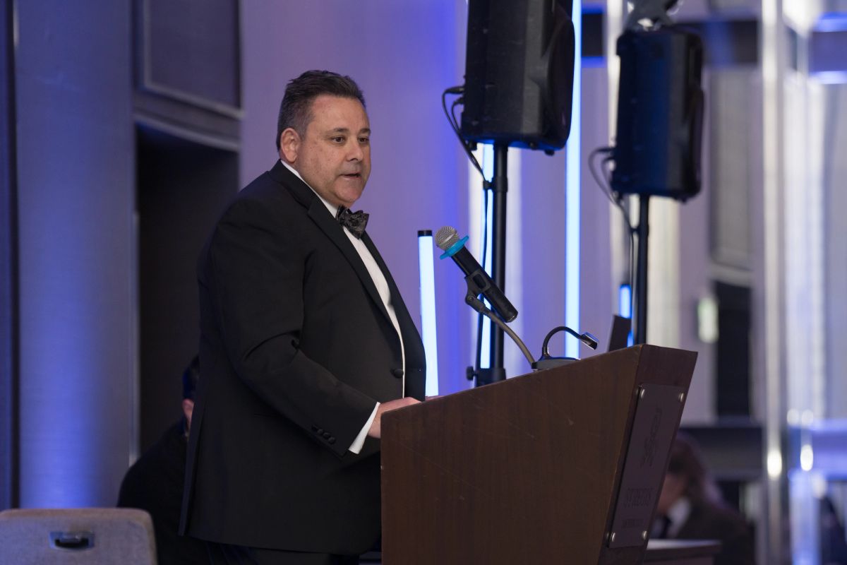 Speaker in black tuxedo presenting at podium with microphone during formal gala event with blue lighting