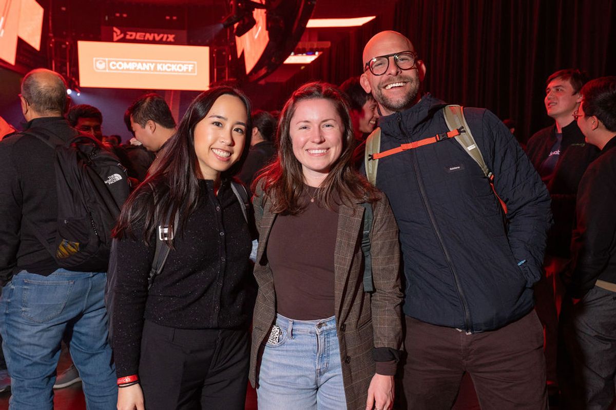 Three conference attendees pose together smiling at an indoor networking event with red lighting and signage.