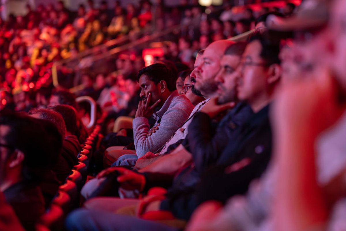 Conference attendees seated in auditorium listening to presentation under red stage lighting