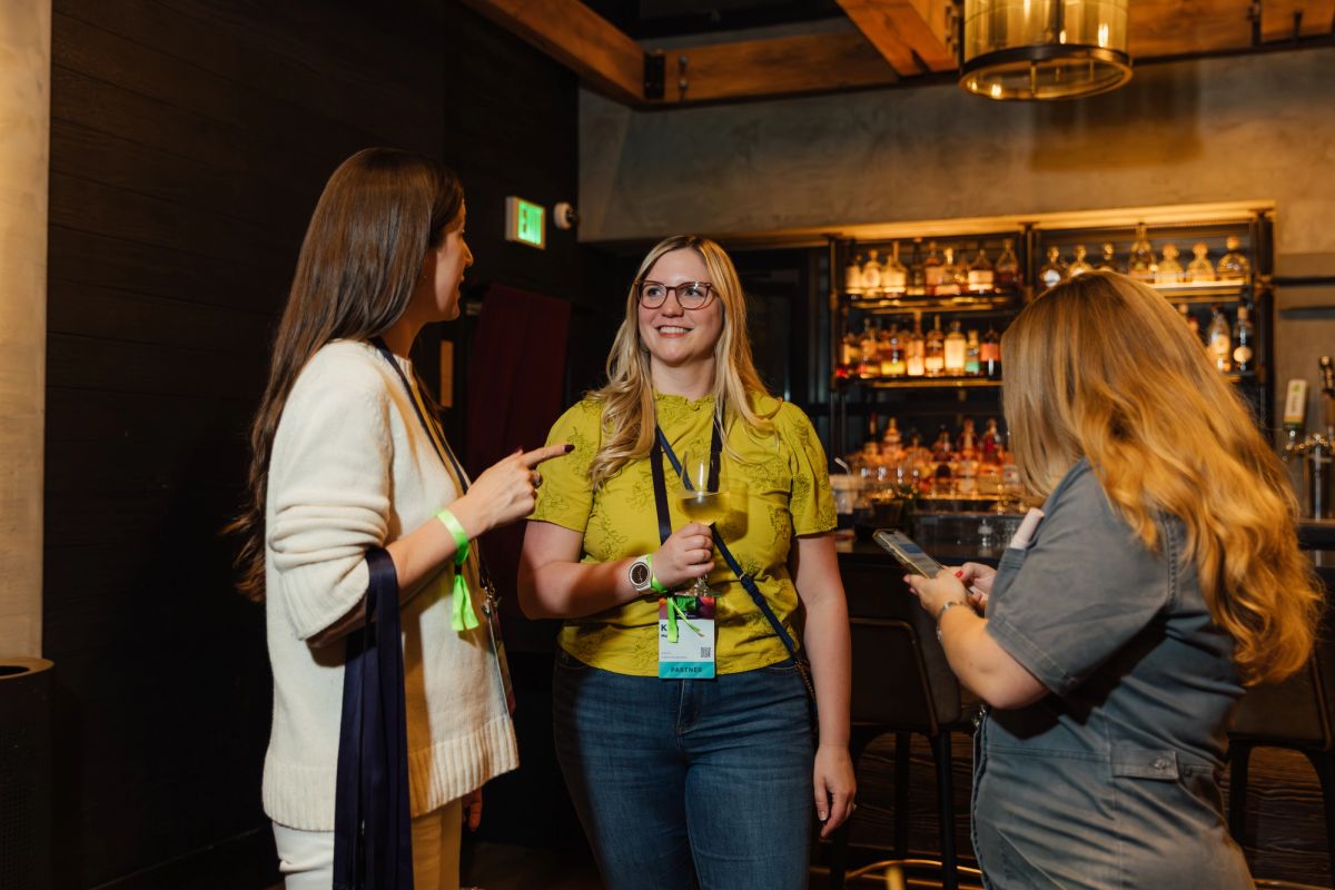 Three women networking at a conference event in an upscale restaurant setting with exposed beams and bar.