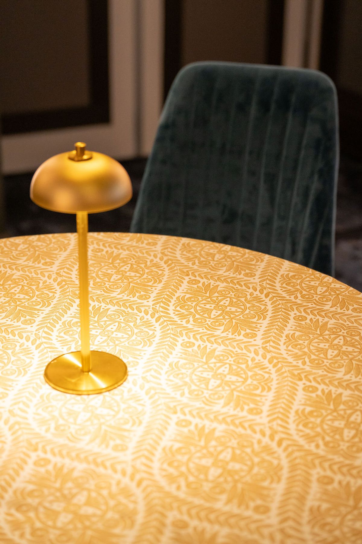 Golden table lamp on patterned yellow tablecloth with teal upholstered chair in background at reception.