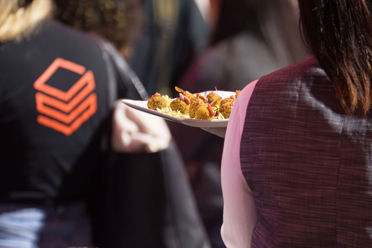 Person in burgundy jacket holds plate of appetizers at conference, attendee in background wears shirt with.