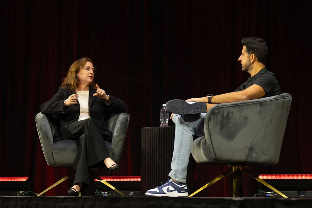 Two people in conversation on stage, seated in gray armchairs with red lighting, one gesturing while speaking