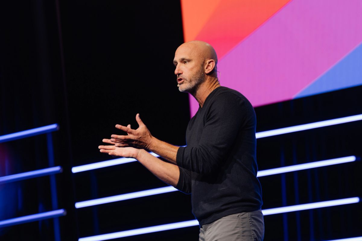 Bald speaker in black shirt gesturing with hands on stage with colorful backdrop and blue lighting