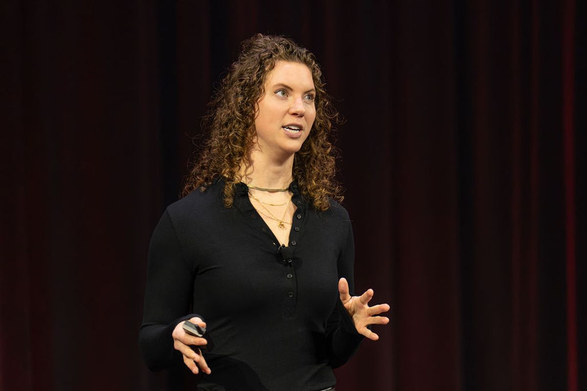 Speaker with curly hair presenting on stage against dark backdrop with red curtains