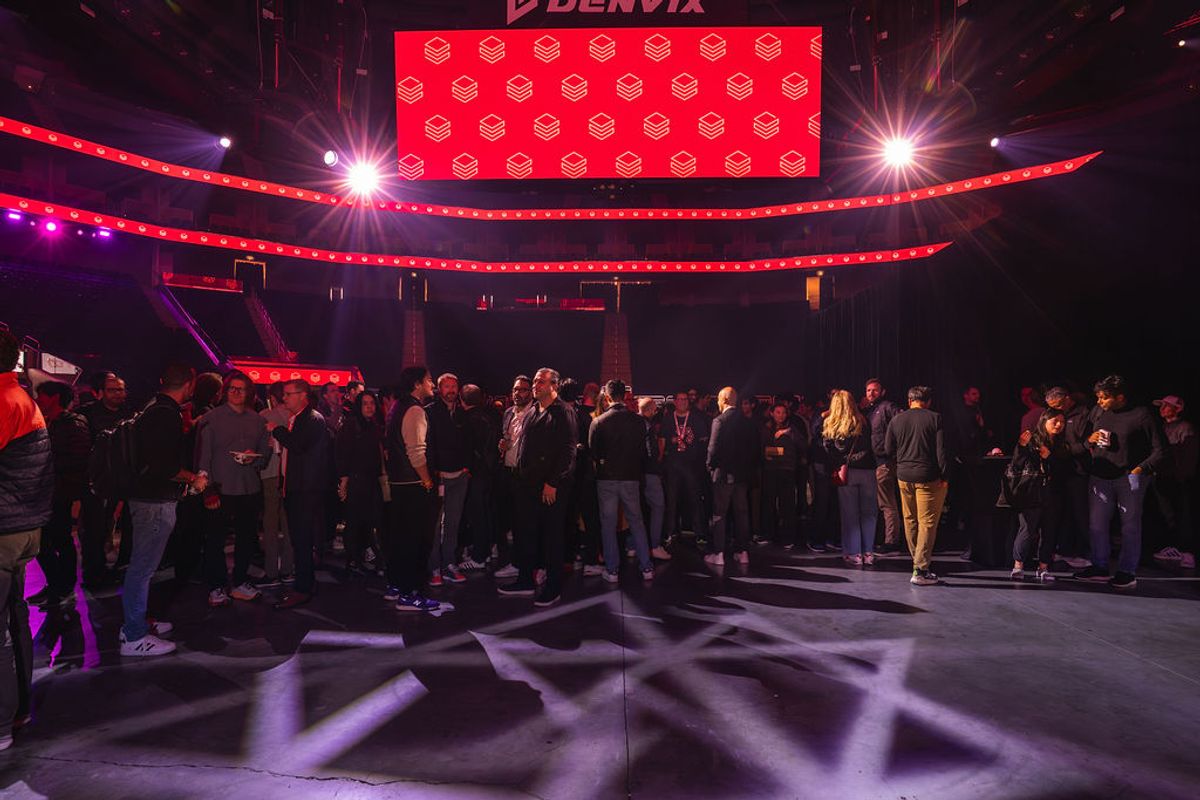 Conference attendees networking in arena venue with red lighting and large screen displaying logo pattern