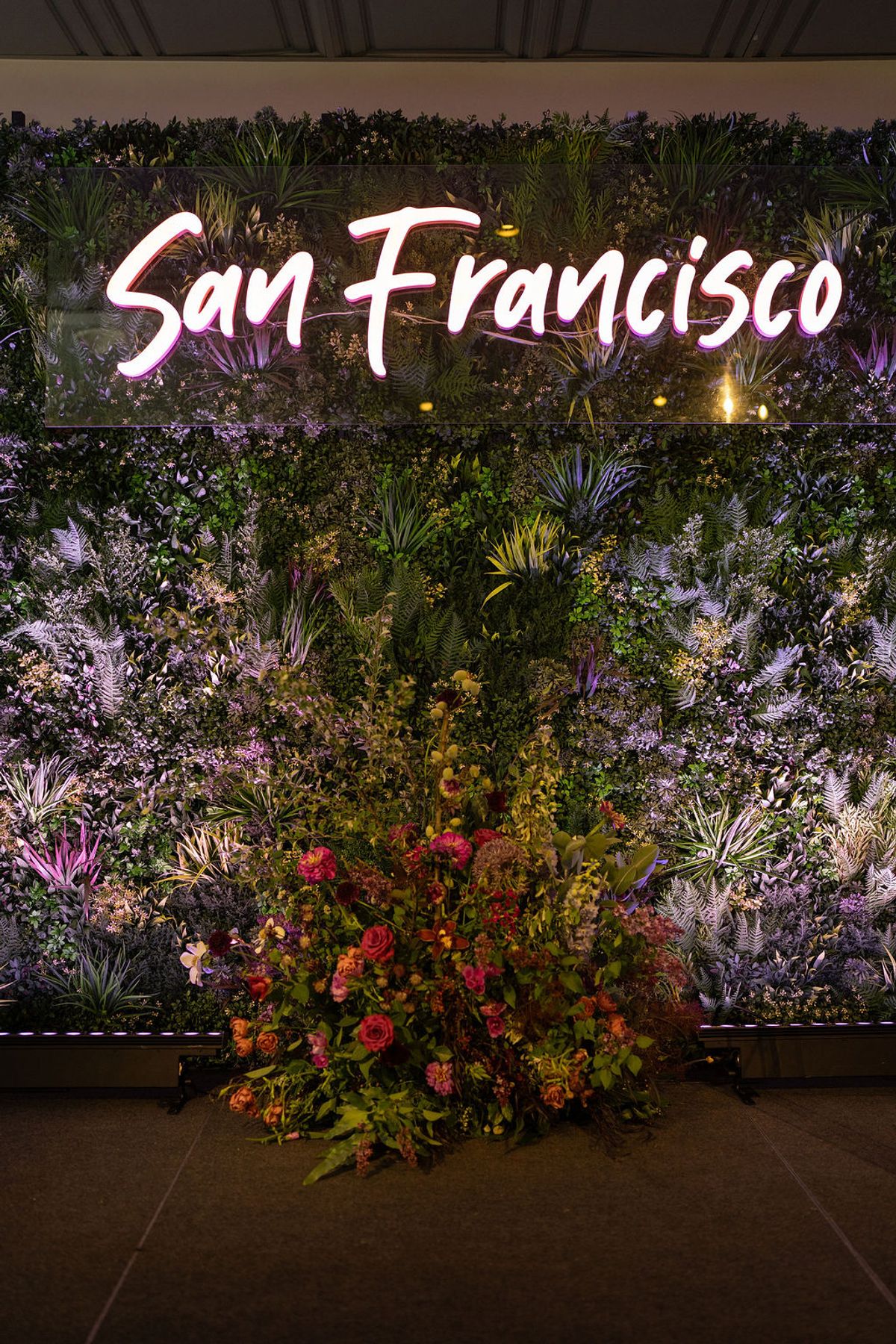 Illuminated "San Francisco" sign on lush green plant wall with colorful floral arrangement at base