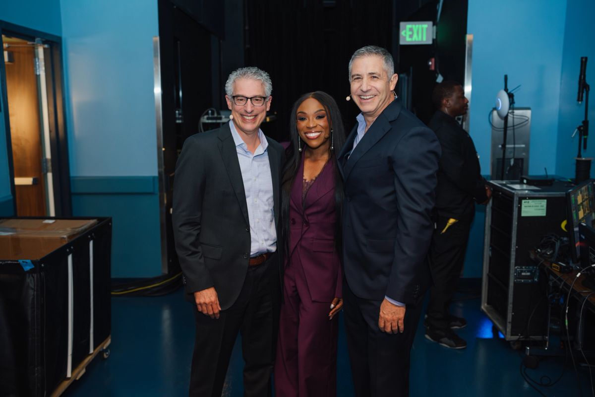 Three people pose together in a blue-lit theater backstage area with equipment and an exit sign visible.