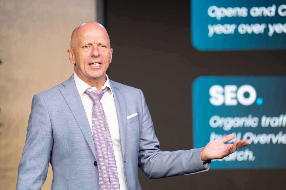 Bald executive in gray suit presenting at conference, gesturing toward blue screen displaying SEO content