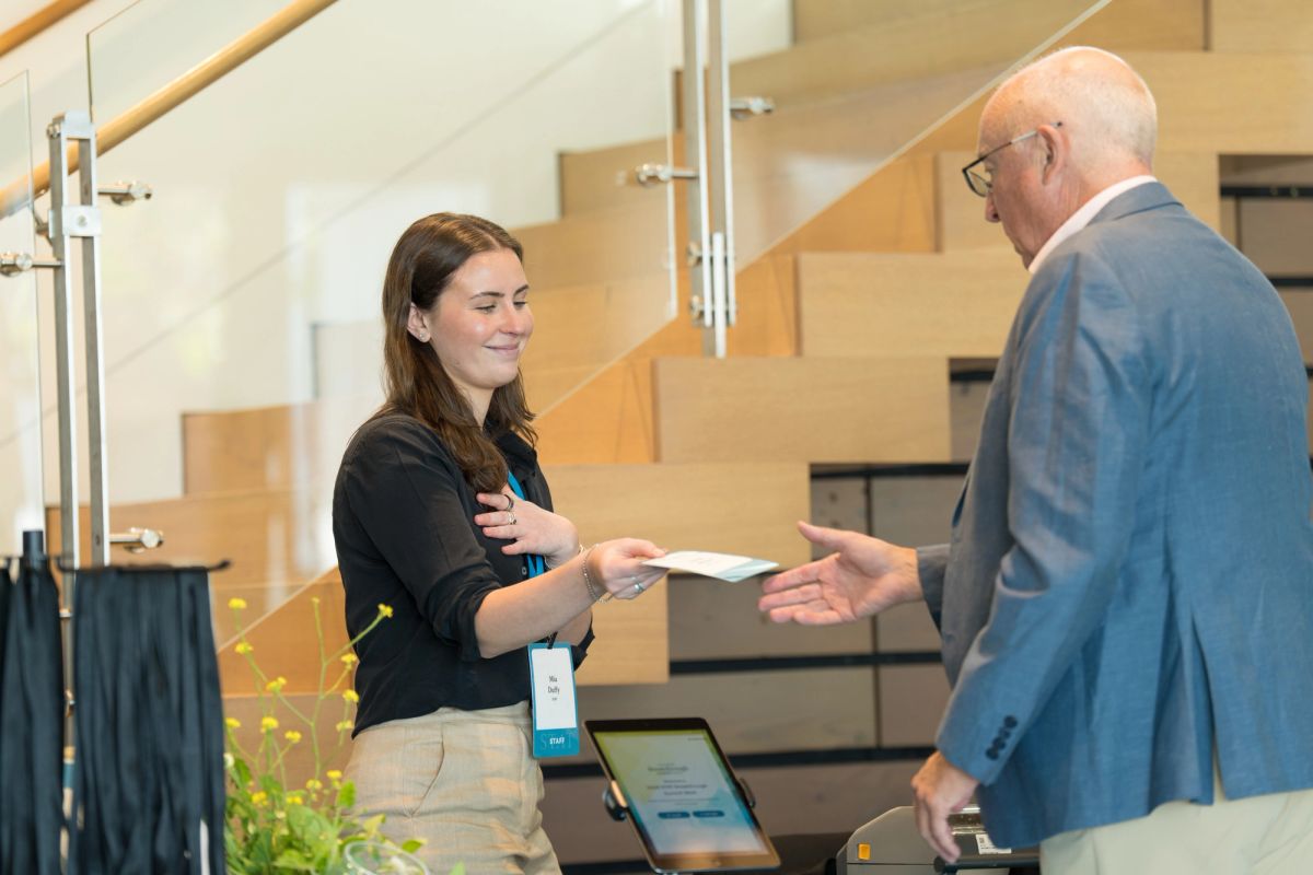 Woman at registration desk hands materials to man in suit at conference venue with modern staircase