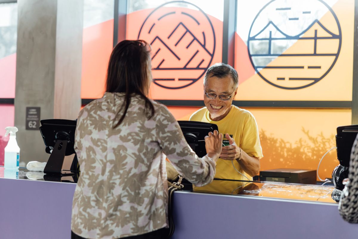 Two people at a purple registration table, one in yellow shirt smiling while holding phone, colorful wall art.