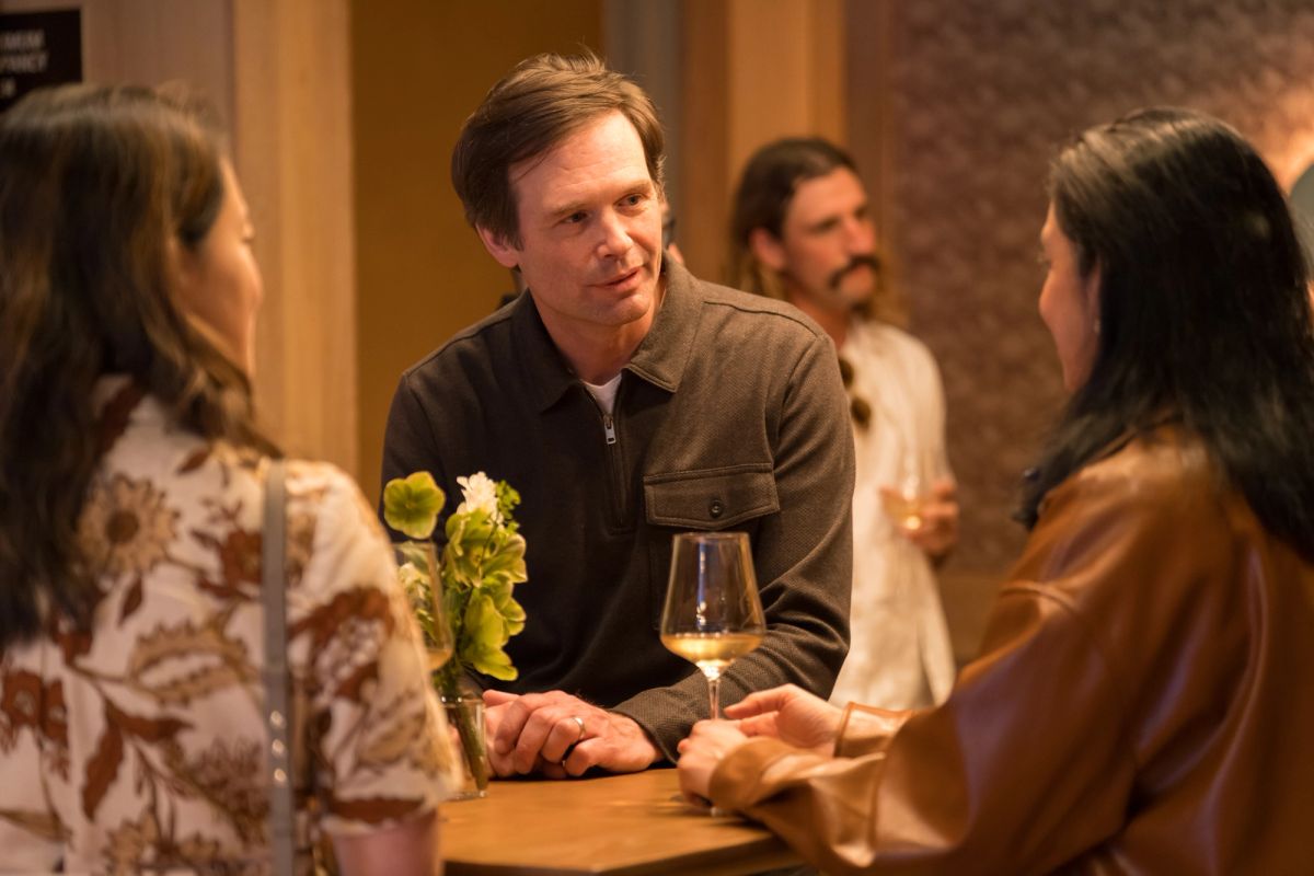 Three people in conversation at a restaurant table with wine glasses and flowers, another person visible in.