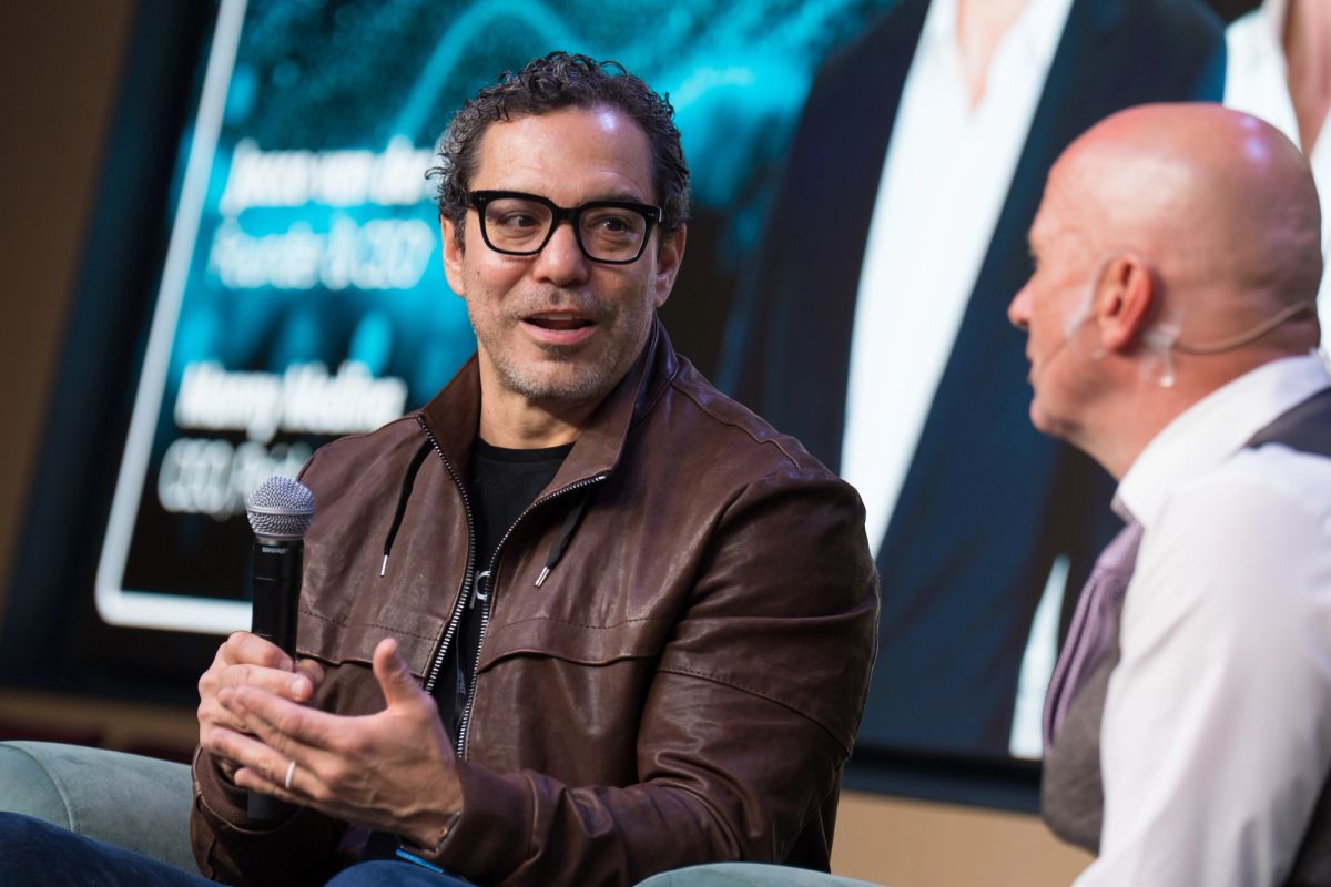 Speaker with glasses and brown leather jacket holds microphone during panel discussion with bald interviewer