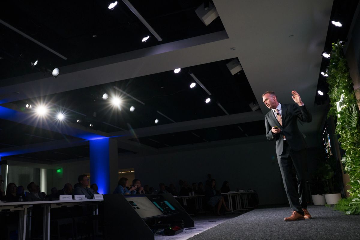 Speaker presenting on stage to seated audience in modern conference venue with blue lighting and exposed.