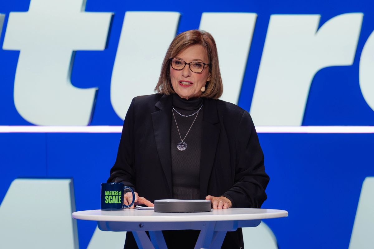 Speaker in black blazer and glasses at white table on blue backdrop stage at conference