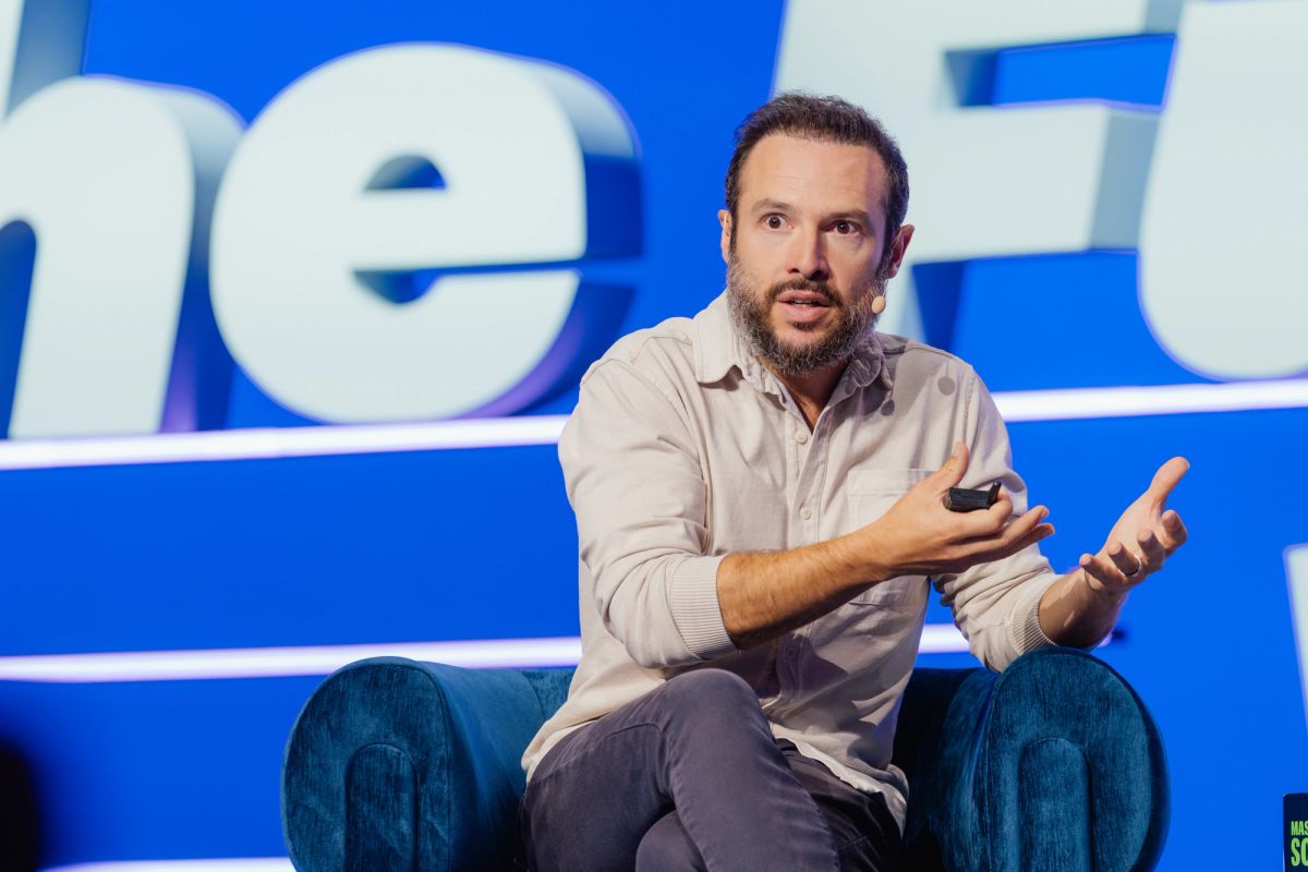 Speaker with beard gesturing while seated on blue chair at conference with blue backdrop