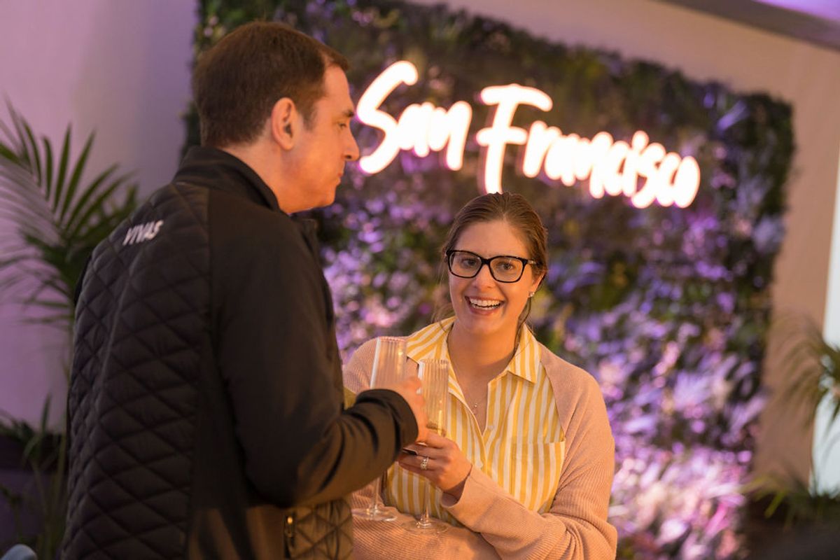 Two attendees networking at reception, man in black jacket interviewing woman with glasses holding drink, San.