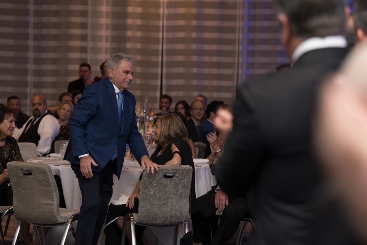 Man in blue suit standing and leaning on chair at formal dinner event with seated guests at round tables