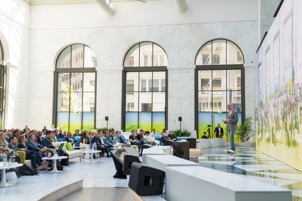 Speaker addresses seated audience in a bright, modern atrium with tall arched windows.