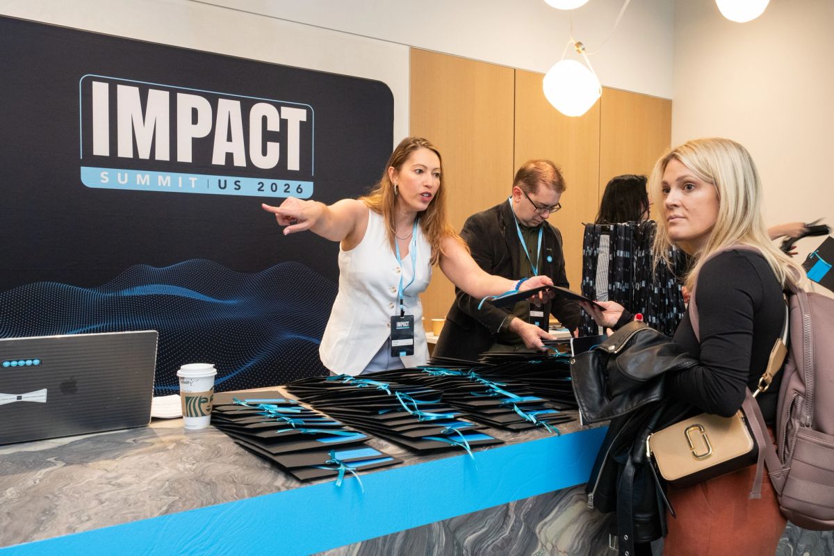 Conference staff member at registration table pointing and assisting attendee at IMPACT Summit US 2026 event